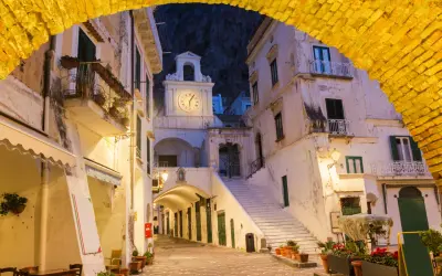 Un vicolo illuminato ad Atrani, con un arco giallo in primo piano e un campanile con orologio sullo sfondo.
