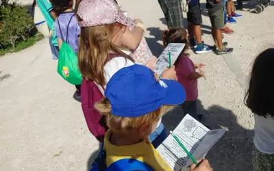 Niños con gorras y mochilas, algunos con folletos, explorando el **parque de Valdebebas** en un día soleado.