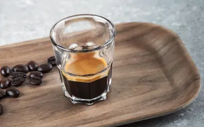 En liten shot med espresso, med crema, bredvid kaffebönor på en träbricka. En perfekt start på dagen, en stark espresso vs. vanligt kaffe.