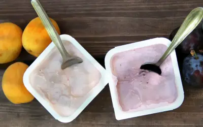 Dwa kubeczki jogurtu owocowego z łyżeczkami, obok morele i śliwki na drewnianym tle.