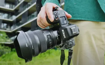 Dłoń trzyma aparat Nikon Z8 z obiektywem. W tle nowoczesny budynek.