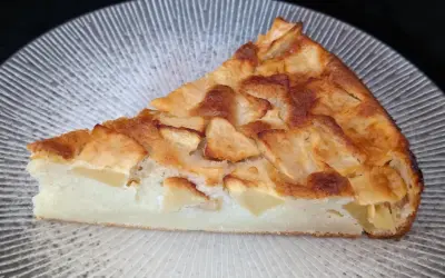 Tranche de clafoutis aux pommes dorée et moelleuse, garnie de morceaux de fruits fondants, sur une assiette texturée.