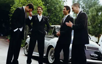 Quattro uomini in abito scuro, perfetti per un matrimonio, ridono vicino a un'auto d'epoca.