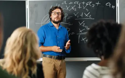 Nauczyciel w niebieskiej koszuli tłumaczy matematykę przy tablicy. Czy nauczyciel jest funkcjonariuszem publicznym? To pytanie nurtuje wielu.