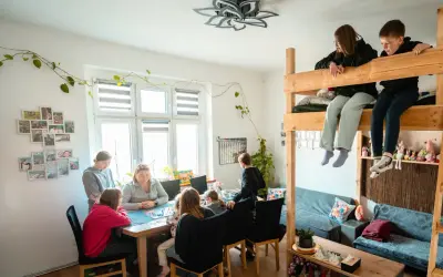 Rodzina gra w planszówkę w domu pomocy społecznej "Przystań". Dzieci siedzą na łóżku piętrowym, obserwując grę.