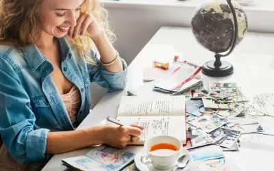 Kobieta pisze esej po angielsku w notatniku, otoczona pamiątkami z podróży: globusem, biletami, zdjęciami i paszportem.