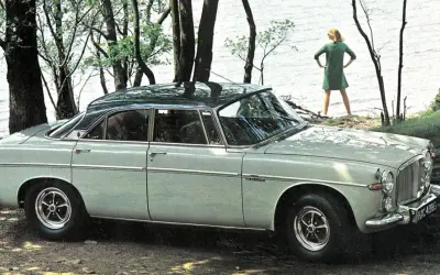 En ljusgrå Rover P5 står parkerad vid en sjö. En kvinna i grön klänning står vid vattenbrynet.