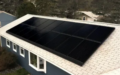 Paneles negros de una aplicacion placas solares cubren el tejado de una casa azul, rodeada de árboles.