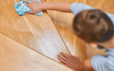 Une main nettoie un escalier en bois verni encrassé avec un chiffon bleu à carreaux.