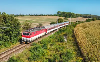 Czerwono-biały pociąg słowacki mija pole kukurydzy i zielone wzgórza w słoneczny dzień.