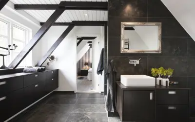 Baño moderno con vigas oscuras y suelo gris. El lavabo sobre encimera y los muebles oscuros contrastan con las paredes de baldosas oscuras. Se ve un dormitorio al fondo.