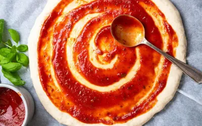 Pizzaboden mit selbstgemachter Pizzasauce bestreichen. Frisches Basilikum und ein Löffel mit Sauce daneben.