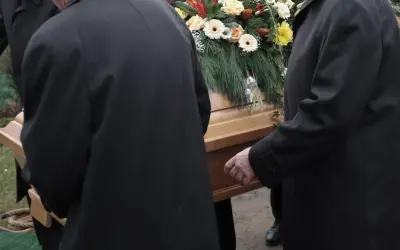 Mężczyźni w czarnych płaszczach niosą trumnę ozdobioną kwiatami. Kondukt pogrzebowy w żałobnej ciszy.