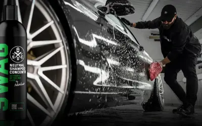 Profesjonalna konserwacja samochodu w pianie. Mężczyzna myje czarne auto gąbką, obok butelka szamponu SWAG.