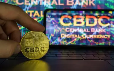 Złota moneta z napisem CBDC w dłoni na tle ekranu z wykresem i napisem "Central Bank Digital Currency". To przyszłość finansów, ale cbdc co to?