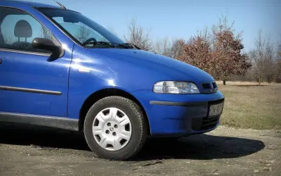 Błękitny Fiat Albea stoi na polnej drodze, w tle jesienne drzewa.