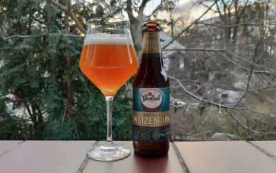 Szklanka z bursztynowym piwem Grolsch Royale Weizen IPA i butelka obok, na tle drzew i nieba.