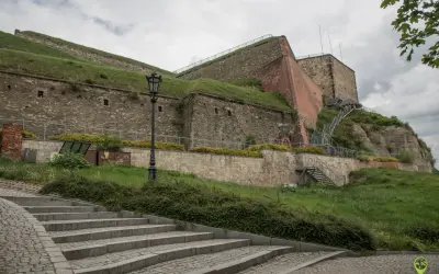 Kamienne schody prowadzą do potężnej twierdzy, jednej z głównych atrakcji Kłodzka. Widać metalowe schody i zielone zbocza.