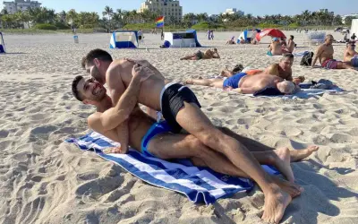 Two men embrace on a towel at Haulover Beach, a popular spot for gay beachgoers. Other sunbathers relax on the sand in the background.