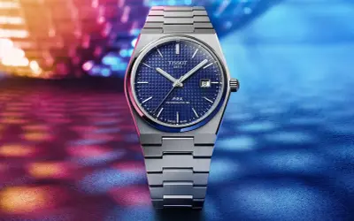Reloj Tissot 1853 PRX Powermatic 80 con esfera azul texturizada y brazalete metálico, sobre un fondo vibrante de luces. Descubre su tissot 1853 precio.