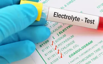 Rękawiczka w niebieskiej rękawiczce trzyma probówkę z napisem "Electrolyte - Test". Obok widać formularz z badaniami, w tym "COAGULOGRAM".