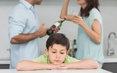 Smutny chłopiec opiera głowę na rękach, podczas gdy rodzice w tle piją alkohol. Obraz symbolizuje trudne dzieciństwo i problemy związane z dda.