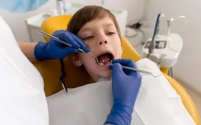 Chłopiec przechodzi leczenie amputacyjne zębów mlecznych. Dentysta w niebieskich rękawiczkach bada jego ząbki.
