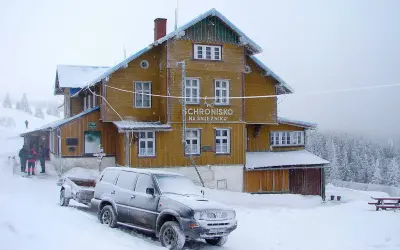 Schronisko na Śnieżniku w zimowej scenerii. Samochód z przyczepą i turyści przy wejściu. Masyw Śnieżnika i jego szlaki w śniegu.