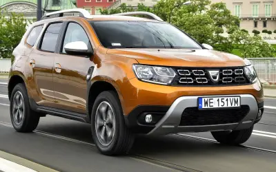 Pomarańczowy Dacia Duster 2 jedzie po moście, za nim widać budynki i drzewa.