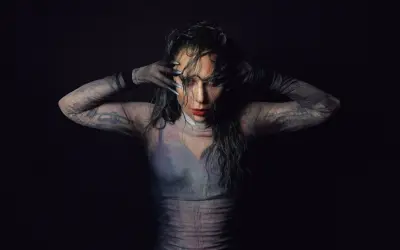 Lady Gaga w siateczkowej bluzce z długimi rękawami i tatuażami, zakrywająca twarz dłońmi.