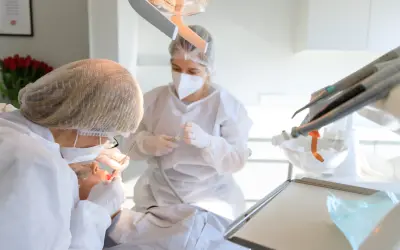 Dentystka w masce i fartuchu wykonuje zabieg kiretażu u pacjentki. W tle widać sprzęt stomatologiczny.
