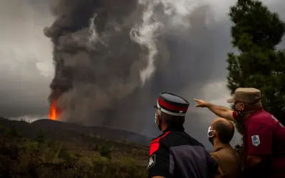 Trzej mężczyźni w maskach obserwują potężną erupcję wulkanu na La Palmie, z której wydobywa się lawa i dym. Obraz przypomina o zagrożeniu tsunami.