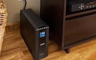 Czarny UPS APC stoi na drewnianej podłodze obok mebla. Jaki UPS, chroni sprzęt przed zanikiem prądu.
