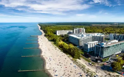Nowoczesny hotel nad samym morzem, z widokiem na piaszczystą plażę pełną wypoczywających ludzi i turkusowe morze.