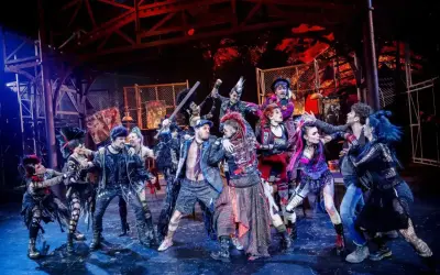 Grupa punkowych artystów na scenie, gotowa do występu. Ich stroje i fryzury krzyczą "we will rock you tłumaczenie" w każdym detalu.