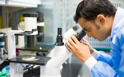 Mężczyzna w niebieskim fartuchu laboratoryjnym patrzy przez mikroskop.
