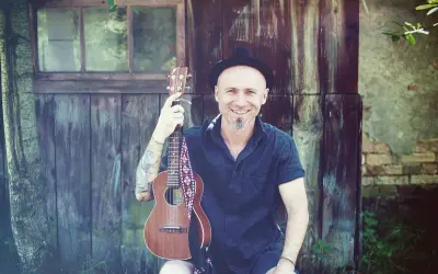 Uśmiechnięty mężczyzna w kapeluszu trzyma ukulele, gotowy do grania nim zajdzie słońce.