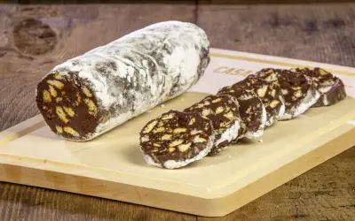 Un rotolo di salame al cioccolato, cosparso di zucchero a velo, è affettato su un tagliere di legno. Pezzi croccanti di biscotto si mescolano al cioccolato fondente.