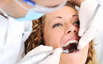 Kobieta u dentysty, zastanawia się ile kosztuje plomba prywatnie. Dentysta w rękawiczkach używa wiertła i lusterka.