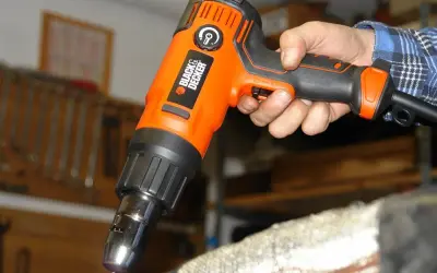 Pomarańczowa opalarka Black & Decker w dłoni, gotowa do pracy. W tle warsztat z narzędziami.