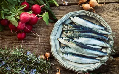 Sardine fresche su piatto d'argento, contornate da ravanelli, rosmarino e cipolline. Un pasto che ricorda una tabella alimenti ricchi di calcio e vitamina D.