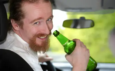 Uśmiechnięty mężczyzna z rudą brodą trzyma zieloną butelkę piwa w samochodzie. Mówi, że piwo to nie alkohol, ale jazda z nim jest niebezpieczna.
