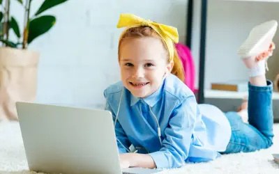 Uśmiechnięta dziewczynka w żółtej opasce i niebieskiej koszuli leży na dywanie z laptopem. Słucha muzyki przez słuchawki, jak wiele dzieci.