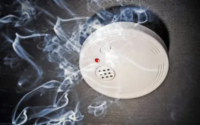 Ein weißer rauchmelder oder feuermelder mit roter LED auf dunklem Grund, umgeben von aufsteigendem Rauch.