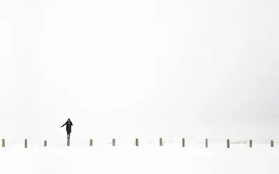 Osoba w czarnym płaszczu balansuje na słupku w śnieżnym krajobrazie. Minimalistyczna, high key fotografia.