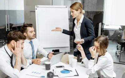En kvinna gestikulerar mot en whiteboard medan kollegor ser utmattade ut. En känsla av passiv aggressiv kommunikation genomsyrar mötet.