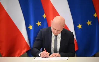 Łysy mężczyzna w garniturze podpisuje dokument na tle flag Polski i UE. Spodziewaj się niespodziewanego, bo nigdy nie wiadomo, co przyniesie przyszłość.