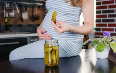 Ciężarna kobieta trzyma w dłoni ogórka kiszonego, obok stoi słoik z ogórkami.