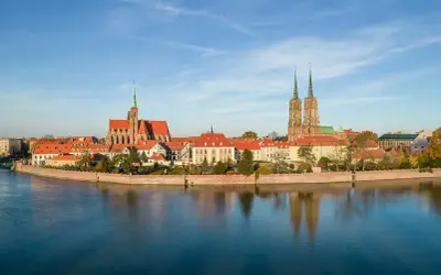 Widok na Ostrów Tumski, serce wrocławskiej starówki, z katedrą i innymi zabytkowymi budynkami odbijającymi się w spokojnej wodzie.