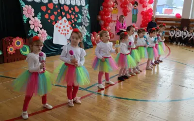 Dzieci w kolorowych spódniczkach śpiewają piosenkę na Dzień Dziecka, trzymając w rękach mikrofony.